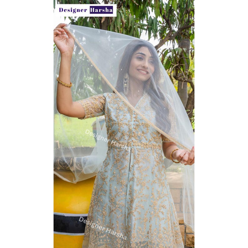 May include: A light blue embroidered dress with a sheer white veil. The dress has a V-neckline and a fitted bodice. The skirt is full and flowing. The veil is draped over the woman's head and shoulders. The dress is decorated with gold embroidery. The image includes the text "Designer Harsha".