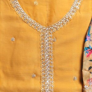 Puede incluir: Una t&uacute;nica de color amarillo mostaza con escote redondo y un panel central vertical adornado con lentejuelas y cuentas plateadas. La tela tiene un acabado texturizado con peque&ntilde;os adornos florales dispersos. El texto DESIGNER HARSHA es visible.