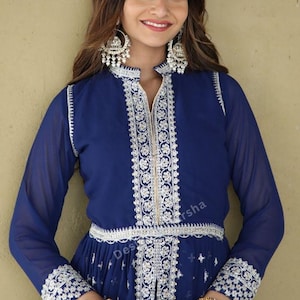 May include: A woman wearing a long, royal blue dress with silver embroidery and a white floral pattern. The dress has a long zipper down the front and a silver belt. The woman is wearing silver earrings and rings.