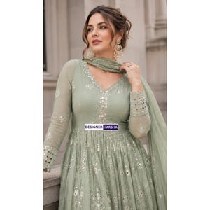 May include: A sage green long-sleeved dress featuring a V-neckline, intricate silver mirror work embroidery, and a matching sheer dupatta. The garment displays the text DESIGNER HARSHA on a small rectangular label centered on the bodice.