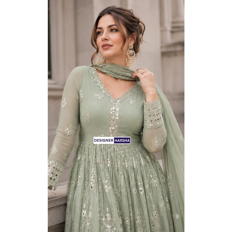 May include: A sage green long-sleeved dress featuring a V-neckline, intricate silver mirror work embroidery, and a matching sheer dupatta. The garment displays the text DESIGNER HARSHA on a small rectangular label centered on the bodice.