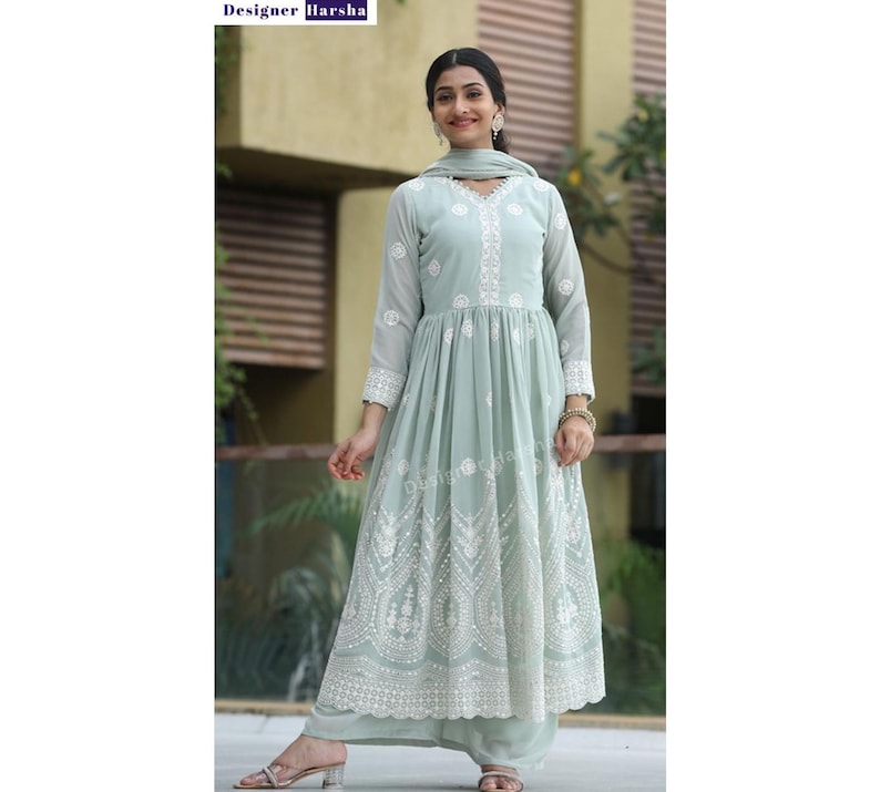 May include: A light green, long-sleeved dress with white embroidery and a matching dupatta. The dress has a gathered skirt and a round neckline. The model is wearing the dress with a pair of silver earrings and a silver bracelet.