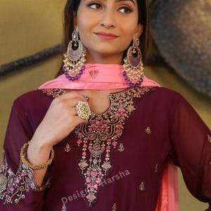 May include: A woman wearing a maroon and pink embroidered dress with a pink dupatta. She is wearing large silver earrings and a ring on her finger. The dress has a floral design and the text "Designer Harsha" is visible on the dress.