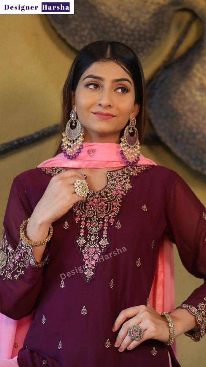 May include: A woman wearing a maroon and pink embroidered dress with a pink dupatta. She is wearing large silver earrings and a ring on her finger. The dress has a floral design and the text "Designer Harsha" is visible on the dress.