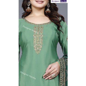 May include: A green salwar kameez with gold embroidery on the neckline. The fabric is a smooth, silky material. The salwar kameez is a traditional Indian garment.