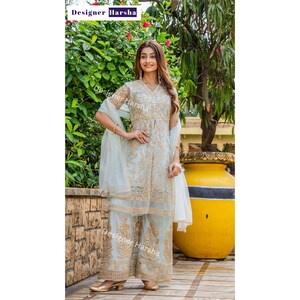 May include: A light blue embroidered dress with gold detailing and a matching dupatta. The dress has a V-neckline and three-quarter sleeves. The model is wearing the dress with palazzo pants and gold sandals. The image has the text "Designer Harsha" in the top left corner and bottom right corner.