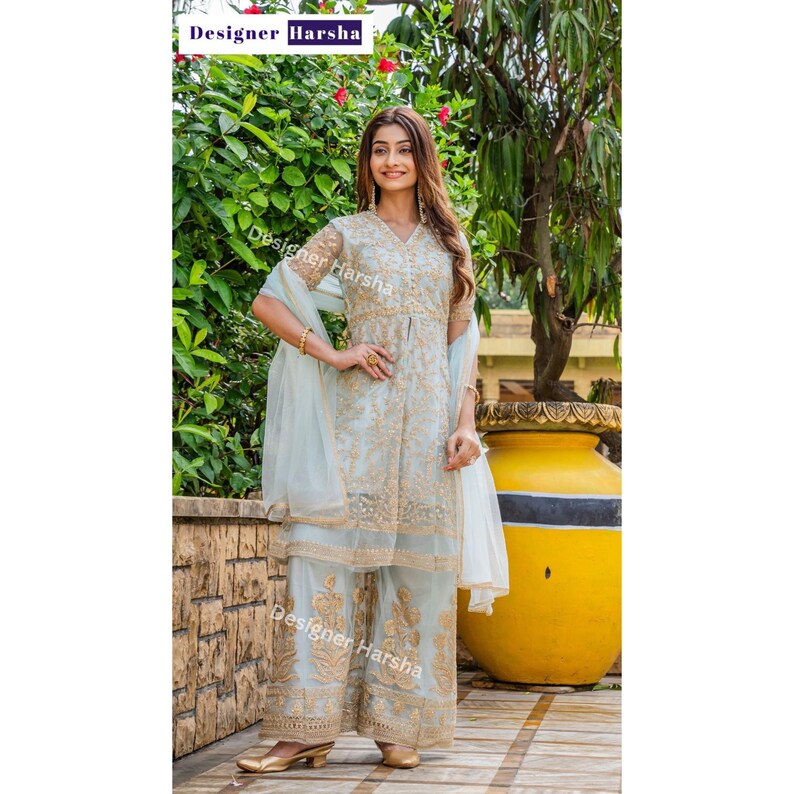 May include: A light blue embroidered dress with gold detailing and a matching dupatta. The dress has a V-neckline and three-quarter sleeves. The model is wearing the dress with palazzo pants and gold sandals. The image has the text "Designer Harsha" in the top left corner and bottom right corner.