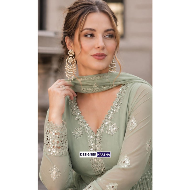May include: A sage green long-sleeved dress featuring intricate silver embroidery and mirror work along the V-neckline and cuffs. The garment includes a matching sheer scarf draped over the shoulder. The text DESIGNER HARSHA is visible.