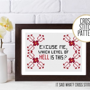 Puede incluir: Patrón de punto de cruz con el texto "Excuse me, which level of hell is this?" y un borde floral rojo. El patrón está enmarcado y se muestra en una pared de ladrillo blanco.