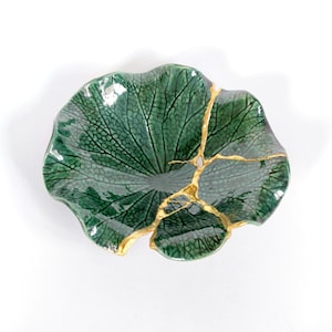 Könnte beinhalten: Eine grüne, keramikartige, blattförmige Schale mit goldfarbener Kintsugi-Reparatur. Die Schale hat eine strukturierte Oberfläche mit einem Blattadermuster. Die goldenen Akzente heben die Risse hervor.