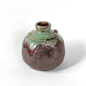 Puede incluir: Un pequeño jarrón de cerámica artesanal con cuerpo redondeado y cuello estrecho. El jarrón presenta una base texturizada de color marrón oscuro, que transiciona a una sección superior verde y marrón rojizo. Dos pequeños elementos decorativos en relieve en el hombro.