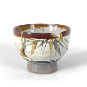 May include: A ceramic bowl featuring a cream-colored body with painted gray and gold leaf bamboo motifs. The rim is accented with a dark brown glaze, and the exterior surface displays gold-filled cracks, a technique known as kintsugi.