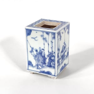 May include: A square ceramic vessel featuring blue and white hand-painted illustrations of figures, bamboo, and landscape elements. The top rim has a blue and white dotted border, and the base is supported by four small feet on the bottom corners.