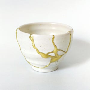 May include: A white ceramic bowl with gold kintsugi repair. The bowl has an irregular shape and a textured surface. The gold lines highlight the cracks, creating a unique aesthetic. The bowl is likely for serving or display. The diameter is approximately 10 cm.