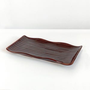 May include: A dark brown ceramic serving platter with a rectangular shape and a wavy edge. The platter has a ridged surface. It is designed for serving food and is set against a white background.