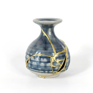 May include: A small, blue ceramic vase with a bulbous body and a narrow neck. The vase features a unique Kintsugi design, with gold-colored lines highlighting the repaired cracks. The base is a light cream color.