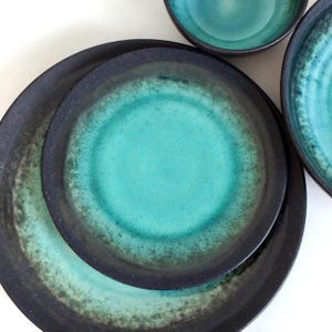 Op de afbeelding: Een set rond keramisch servies met een donkere, matzwarte buitenrand en een levendig turquoise blauw midden. Het glazuur verloopt van diep petrol naar helder cyaan, wat zorgt voor een getextureerd en gespikkeld uiterlijk op de borden en kommen.