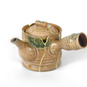 Op de afbeelding: Een keramische theepot met een zijhandvat en een deksel, voorzien van een beige en groene glazuurlaag. De theepot vertoont zichtbare barsten die zijn gerepareerd met goudlak, een techniek genaamd kintsugi, die de breuken op het lichaam accentueert.