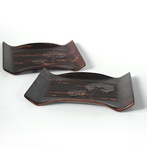 May include: Two dark brown wooden trays with a curved design and carved tree and line details. The trays have a rectangular shape with a slightly raised edge and are likely used for serving or display.
