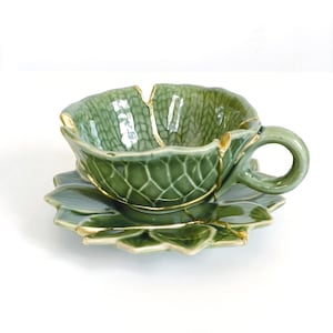 May include: A green ceramic teacup and saucer set. The cup and saucer are leaf-shaped with a textured surface and gold accents. The teacup has a handle and sits on a matching saucer.