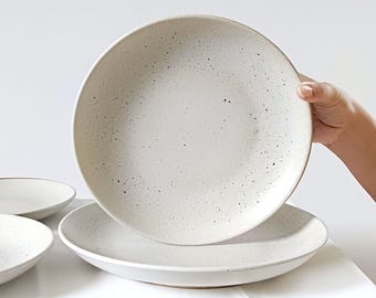 May include: A set of speckled white ceramic plates. One plate is being held up, revealing a similar plate beneath it. The plates have a slightly raised edge and a speckled pattern, with small dark spots. The plates are on a white surface.