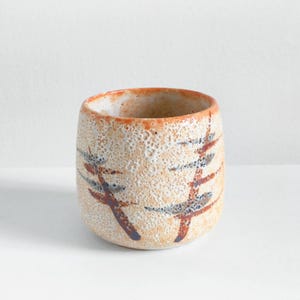 Puede incluir: Una pequeña taza de cerámica texturizada en beige y naranja. Tiene un diseño de ramas de árbol en marrón y azul. El borde es naranja. La taza probablemente se usa para beber. Aproximadamente 8 cm de alto.