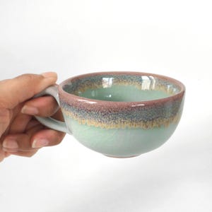 Op de afbeelding: Een keramische mok met een brede, ronde komvorm en een handvat aan de zijkant. De buitenkant heeft een licht muntgroene basis met een opvallende craquelé glazuurafwerking, bekroond door een decoratieve band van gemengd mauve, blauw en goudbruin druipglazuur.