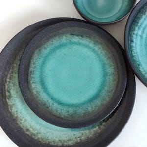 May include: A collection of ceramic tableware in teal and black. The plates and bowls have a textured, glazed finish. The plates are stacked, showing the different sizes and depths of the items. The set is suitable for serving food.