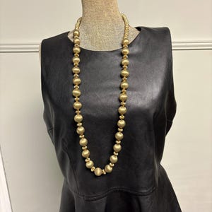May include: A long gold-toned necklace with large, round beads and smaller, textured beads. The necklace is displayed on a mannequin wearing a black sleeveless top. The beads have a matte finish and are evenly spaced.