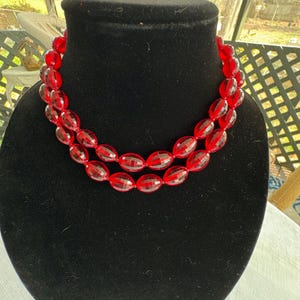 May include: A two-strand red beaded necklace. The beads are oval-shaped with a faceted design, giving them a sparkling appearance. The necklace is displayed on a black velvet bust, highlighting the vibrant red color.