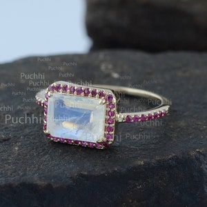 Może przedstawiać: Złoty pierścionek z prostokątnym kamieniem księżycowym osadzonym w kwadratowej ramie z małych różowych kamieni szlachetnych. Pierścionek leży na ciemnoszarej, fakturowanej powierzchni.