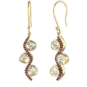 May include: Gold earrings with a curved design. Each earring features three clear, faceted stones and a line of small, red gemstones. The earrings have a hook closure and are displayed against a white background.