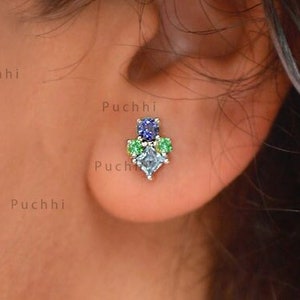 May include: A close-up of a person's ear with a small, silver earring featuring a cluster of blue, green, and clear gemstones.