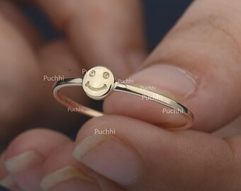 May include: A thin gold ring featuring a small, circular charm with an engraved smiley face design. The charm displays two small dots for eyes and a curved line for a mouth. The word Puchhi is visible multiple times across the metal surface.