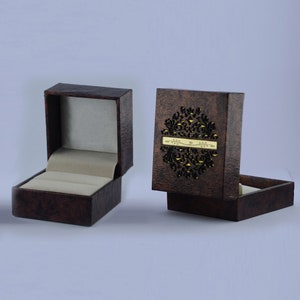 May include: A brown ring box with a decorative cutout design and a gold inscription. The box is open and the inside is lined with a light gray fabric.
