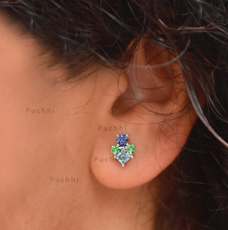 May include: A close-up of a person's ear with a small, silver earring featuring a cluster of blue, green, and clear gemstones.