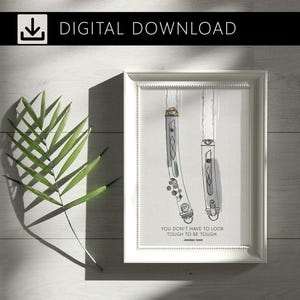 Peut inclure: Une impression encadrée avec une bordure blanche représentant deux illustrations de sabres laser. Les sabres laser sont gris et argentés avec des lames blanches. Le texte "YOU DON'T HAVE TO LOOK TOUGH TO BE TOUGH. AHSOKA TANO" est en dessous. Le texte "DIGITAL DOWNLOAD" est en haut.
