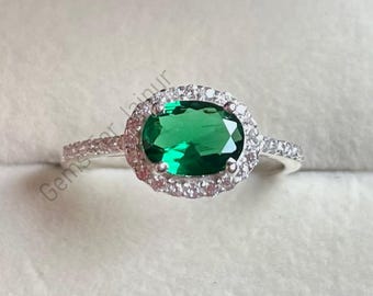 May include: A silver ring featuring a large, oval-cut emerald green gemstone surrounded by a halo of smaller, clear gemstones. The ring's band is also adorned with small, clear gemstones. The ring is set against a soft, off-white background.
