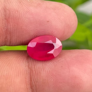 May include: A polished, oval-cut ruby gemstone. The deep red gem is held between fingers, displaying its multiple facets. The ruby's surface reflects light, highlighting its brilliance and colour.