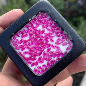 Könnte beinhalten: Ein schwarzes, quadratisches Schmuckkästchen gefüllt mit zahlreichen, marquiseförmigen, leuchtend pinkfarbenen Edelsteinen. Die Edelsteine sind auf einer weißen Oberfläche angeordnet und erzeugen eine lebendige Darstellung. Das Etui wird in einer Hand gehalten, mit unscharfem Hintergrund.