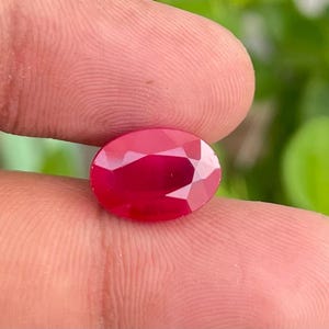 Op de afbeelding: Een gefacetteerde, ovaalvormige robijn edelsteen. De robijn heeft een diepe, verzadigde rode kleur met een briljante glans. De edelsteen wordt tussen twee vingers gehouden, met een vage groene achtergrond.