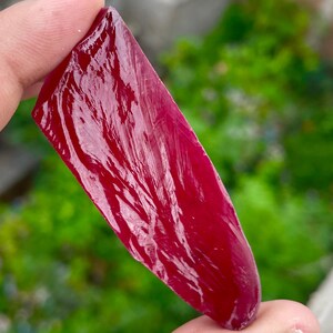 May include: A polished, deep red gemstone with a smooth, reflective surface. The elongated, irregular shape of the stone is held between fingers, with a blurred green background.