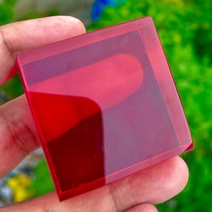 May include: A square-shaped, translucent red glass block with smooth, flat surfaces and sharp, defined edges. The material is clear enough to allow light to pass through, creating a vibrant crimson hue with subtle reflections on the top surface.