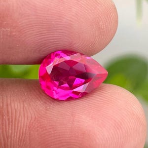 May include: A faceted, pear-shaped, hot pink gemstone. The gem is held between two fingers, with a blurred green and white background. The gem's cut creates many reflective surfaces.