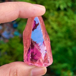 Puede incluir: Un cristal rosa facetado, sostenido entre los dedos. El cristal tiene una cualidad translúcida, con reflejos internos de color azul y púrpura. El fondo es verde borroso, lo que sugiere un entorno al aire libre. La forma del cristal es irregular.