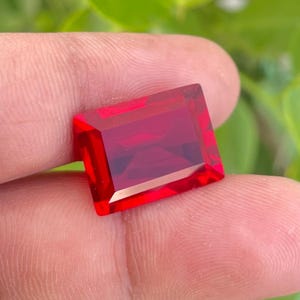 May include: A faceted, rectangular-cut, deep red gemstone. The gem has sharp edges and a polished surface, reflecting light. The stone is held in a hand, with a blurred green background.