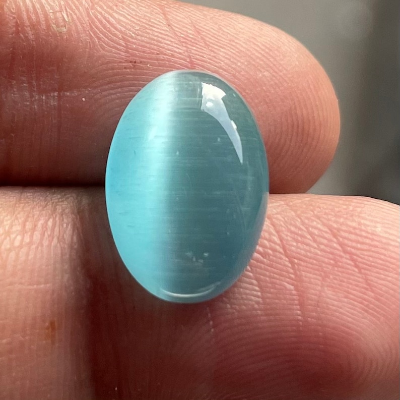 May include: A light blue oval cabochon gemstone with a chatoyant effect. The stone has a smooth, polished surface and a translucent appearance.