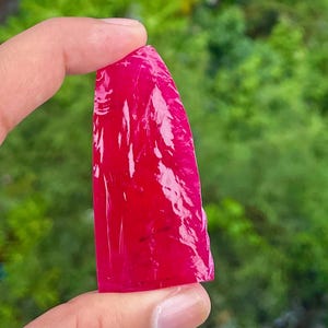May include: A translucent, vibrant magenta-red gemstone with a smooth, polished surface and a slightly irregular, elongated shape. The stone exhibits a glossy, reflective texture that catches the light, showcasing its deep, saturated colour throughout.