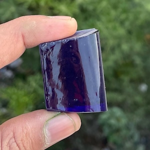 May include: A rectangular, deep blue glass object is held between fingers. The glass has a glossy, reflective surface. The edges are cleanly cut, and the overall shape is a slightly irregular rectangle. The background is blurred green foliage.
