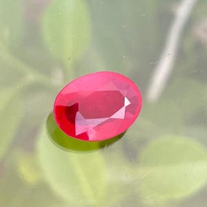 Może przedstawiać: Fasetowany, owalny, ciemnoczerwony kamień szlachetny. Klejnot ma wypolerowaną powierzchnię z wieloma fasetami, które odbijają światło. Kamień jest osadzony na rozmytym zielono-szarym tle, tworząc kontrast, który podkreśla kolor i blask klejnotu.